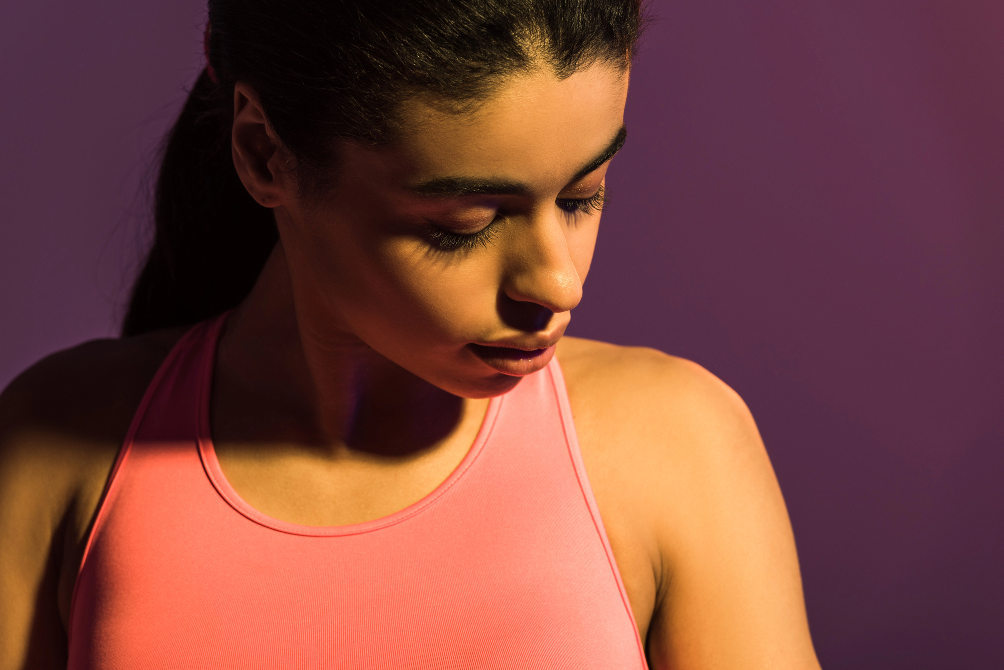 A woman wearing a pink athletic top looks downward with a neutral expression. The background is a soft gradient of purple and pink, creating a serene and focused atmosphere. Her hair is tied back, and the lighting creates gentle shadows on her face and shoulder.