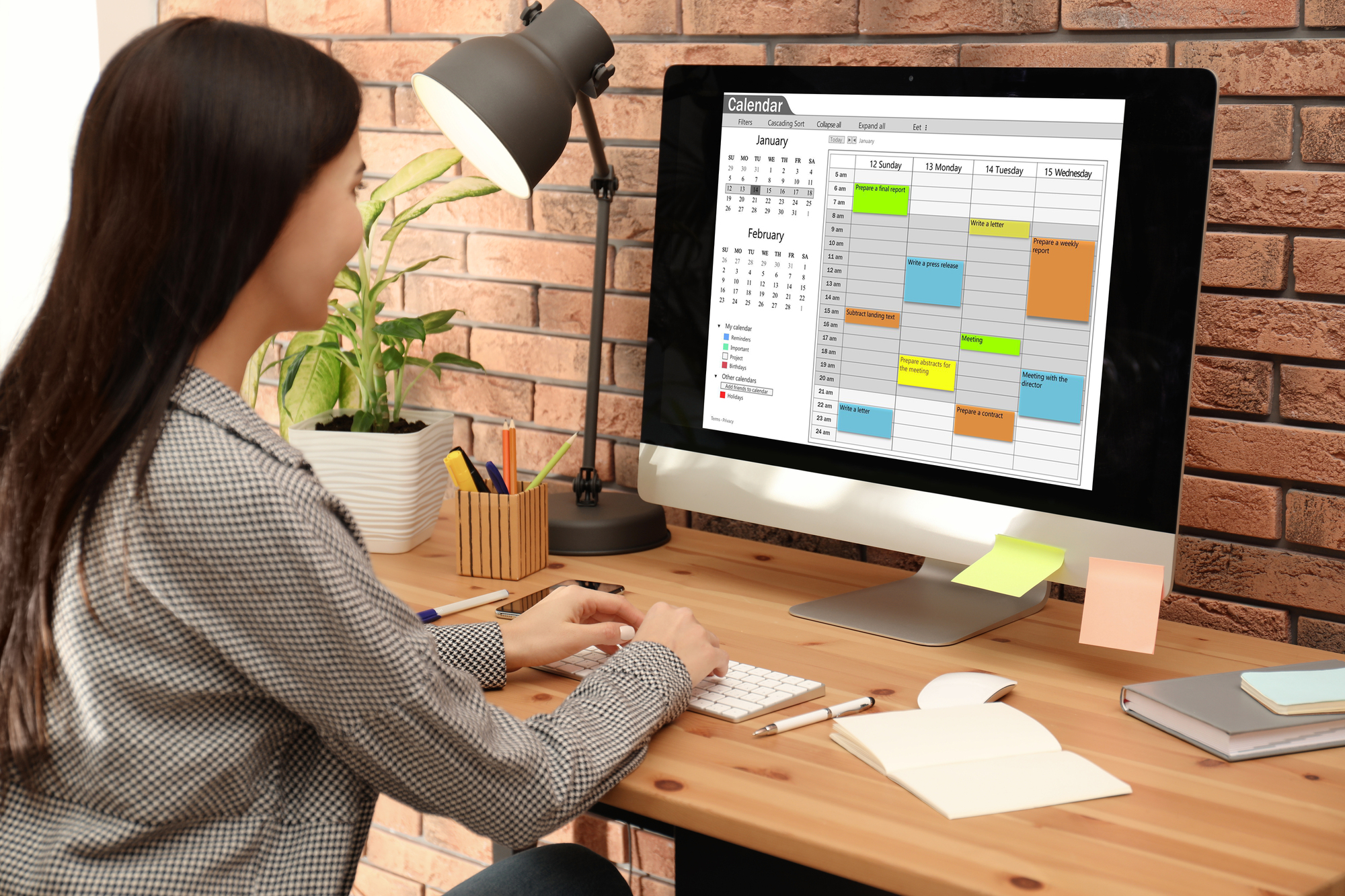 A woman is sitting at a desk, facing a computer displaying a digital calendar with colorful events scheduled. The desk has office supplies, plants, and sticky notes. A lamp is turned on, illuminating the work area. The background features a brick wall.