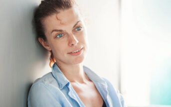 A woman with light skin, brown hair pulled back, and blue eyes is leaning against a light-colored wall. She is wearing a light blue button-up shirt over a black top. The background is softly lit, creating a calm and relaxed atmosphere.