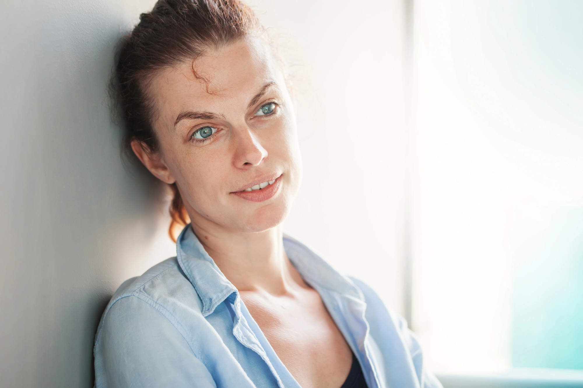 A woman with light skin, brown hair pulled back, and blue eyes is leaning against a light-colored wall. She is wearing a light blue button-up shirt over a black top. The background is softly lit, creating a calm and relaxed atmosphere.