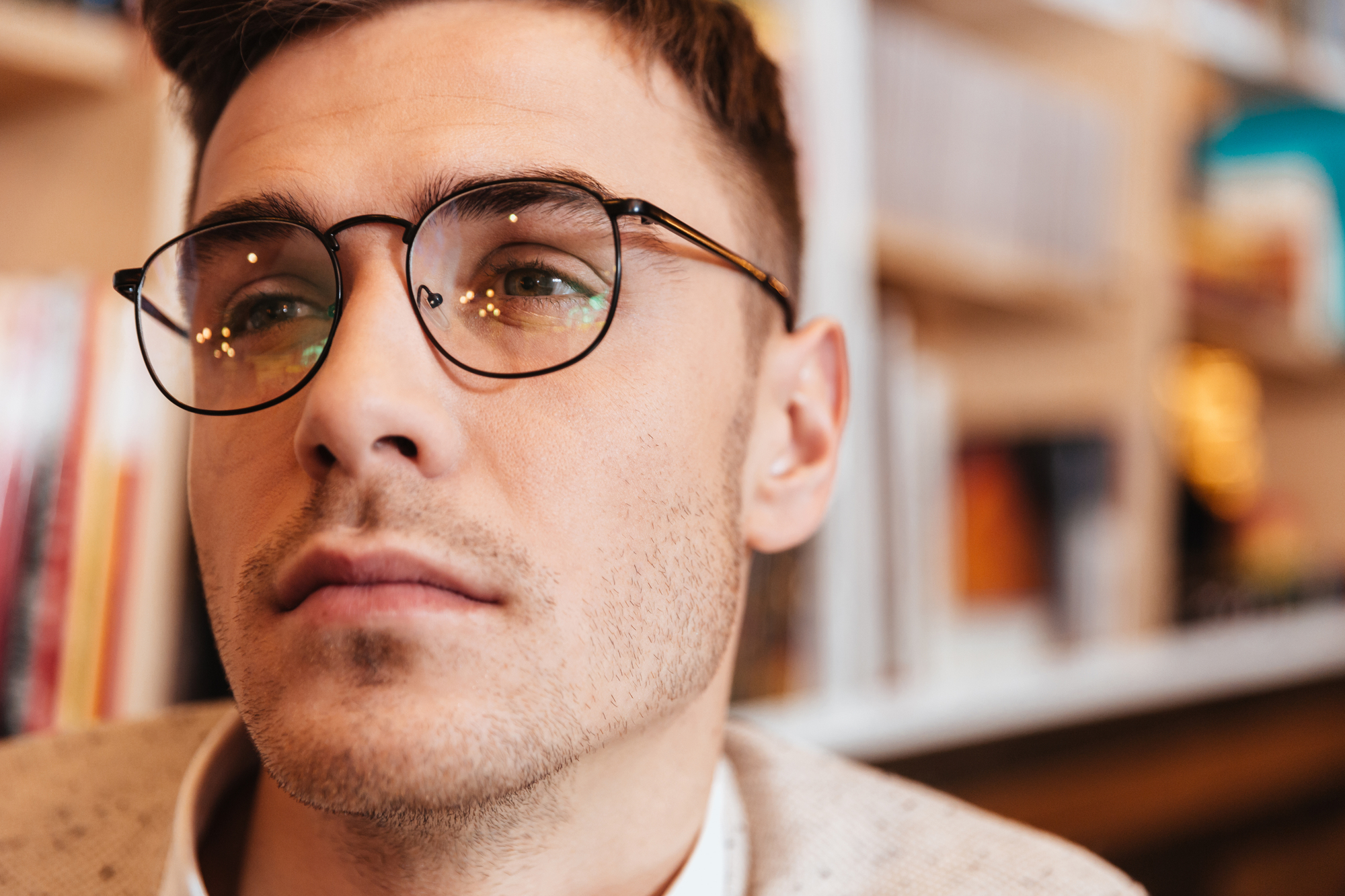 A close-up of a person with short brown hair and glasses, looking into the distance. The background is blurred but appears to be a bookshelf with various books and items. The person has a contemplative expression and wears a light-colored shirt and jacket.