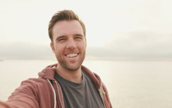 A man with short brown hair and a beard is taking a selfie outdoors. He is wearing a dark T-shirt and a maroon hoodie. He is smiling against a backdrop of a calm, sunlit body of water, with a light, hazy sky above.