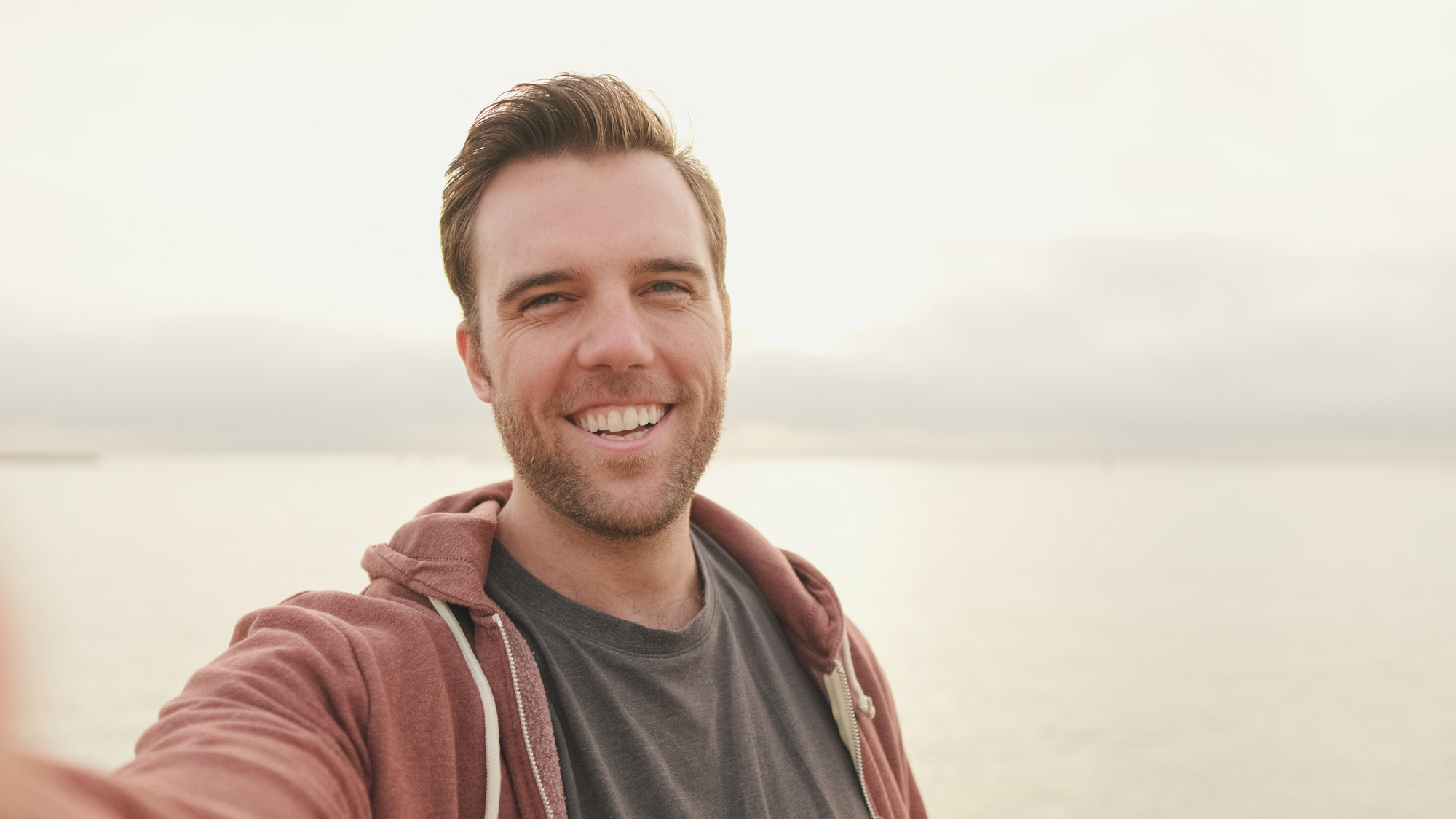 A man with short brown hair and a beard is taking a selfie outdoors. He is wearing a dark T-shirt and a maroon hoodie. He is smiling against a backdrop of a calm, sunlit body of water, with a light, hazy sky above.