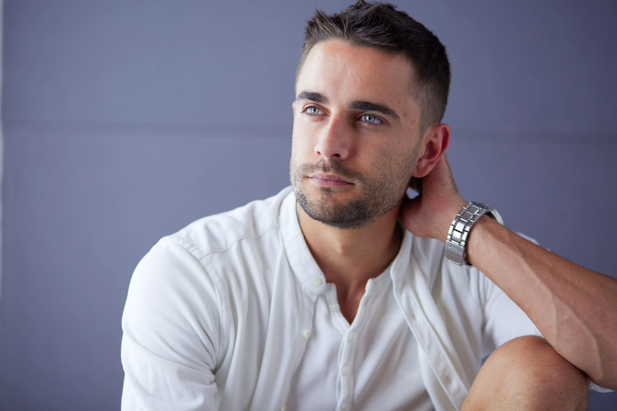 A man with short dark hair and a trimmed beard looks off to the side while touching the back of his neck. He is wearing a white button-up shirt and a silver wristwatch. The background is a plain grey wall.