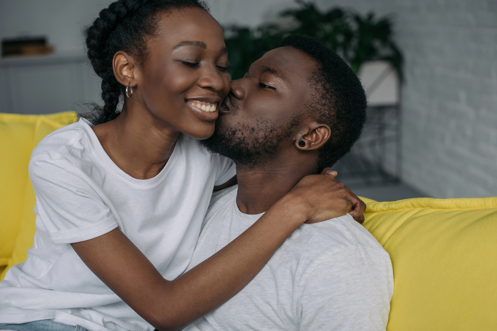 A woman is smiling with her eyes closed while receiving a kiss on the cheek from a man. They are sitting close together on a yellow couch, both wearing white shirts. The background includes blurred indoor plants and a light-colored wall.