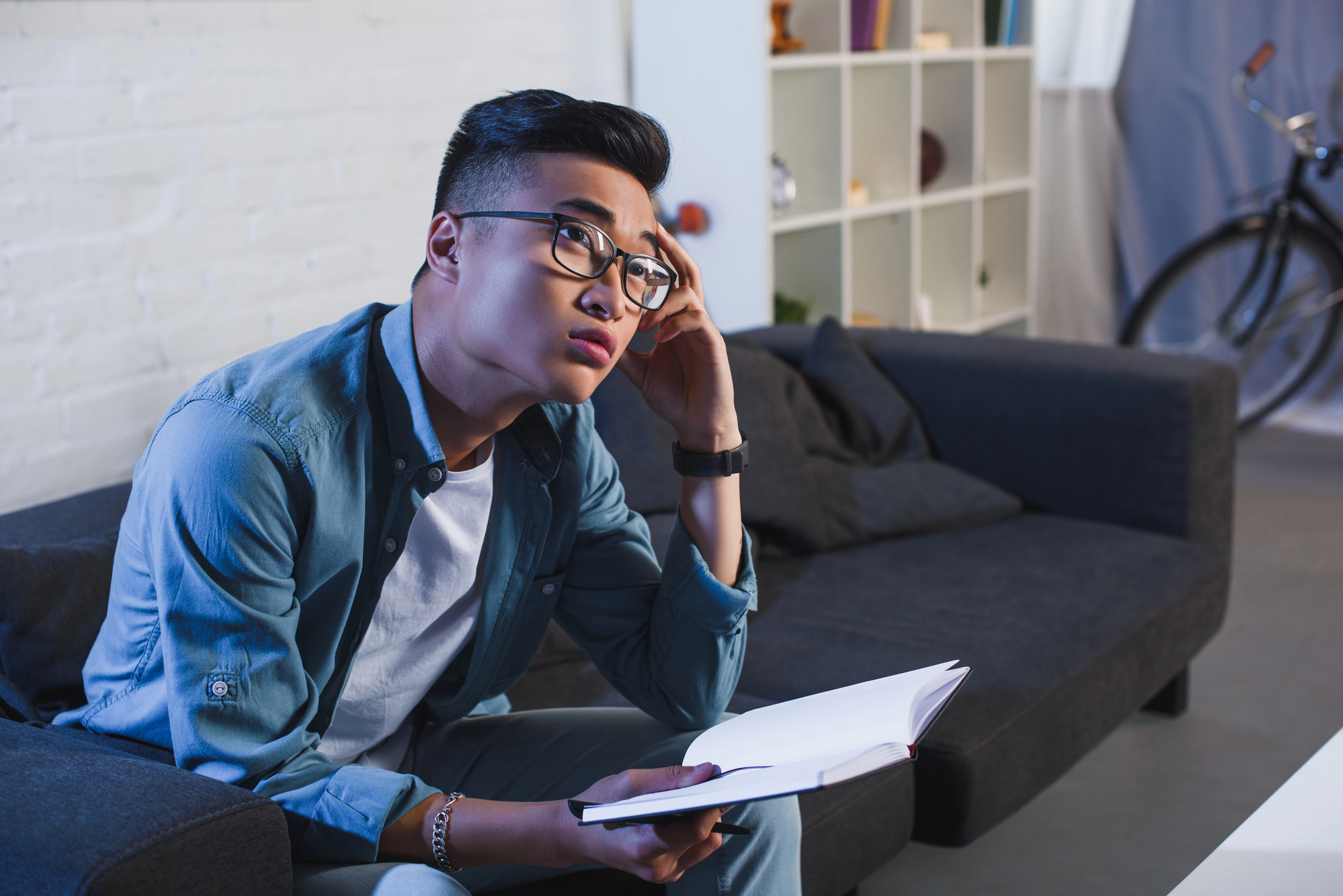 A young man wearing glasses and casual clothes sits on a dark gray couch in a modern living room. He holds an open book and rests his head on his left hand, looking pensive. A bicycle is visible in the background near a white shelving unit.