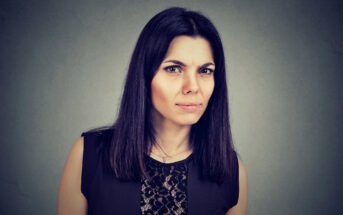 A woman with long dark hair and a serious facial expression is staring directly at the camera. She is wearing a black sleeveless top with a lace pattern on the front. The background is a plain, muted gray.