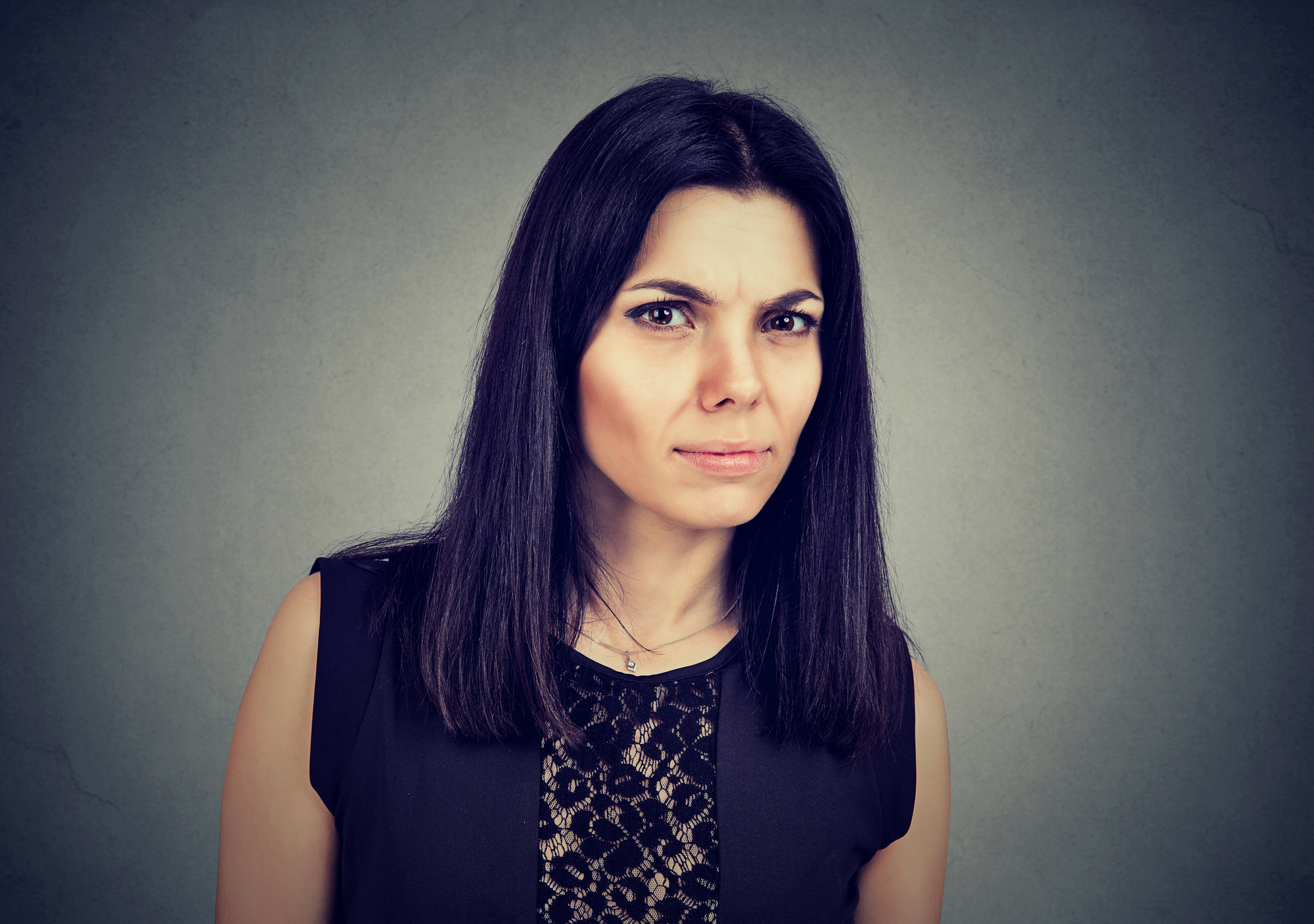 A woman with long dark hair and a serious facial expression is staring directly at the camera. She is wearing a black sleeveless top with a lace pattern on the front. The background is a plain, muted gray.