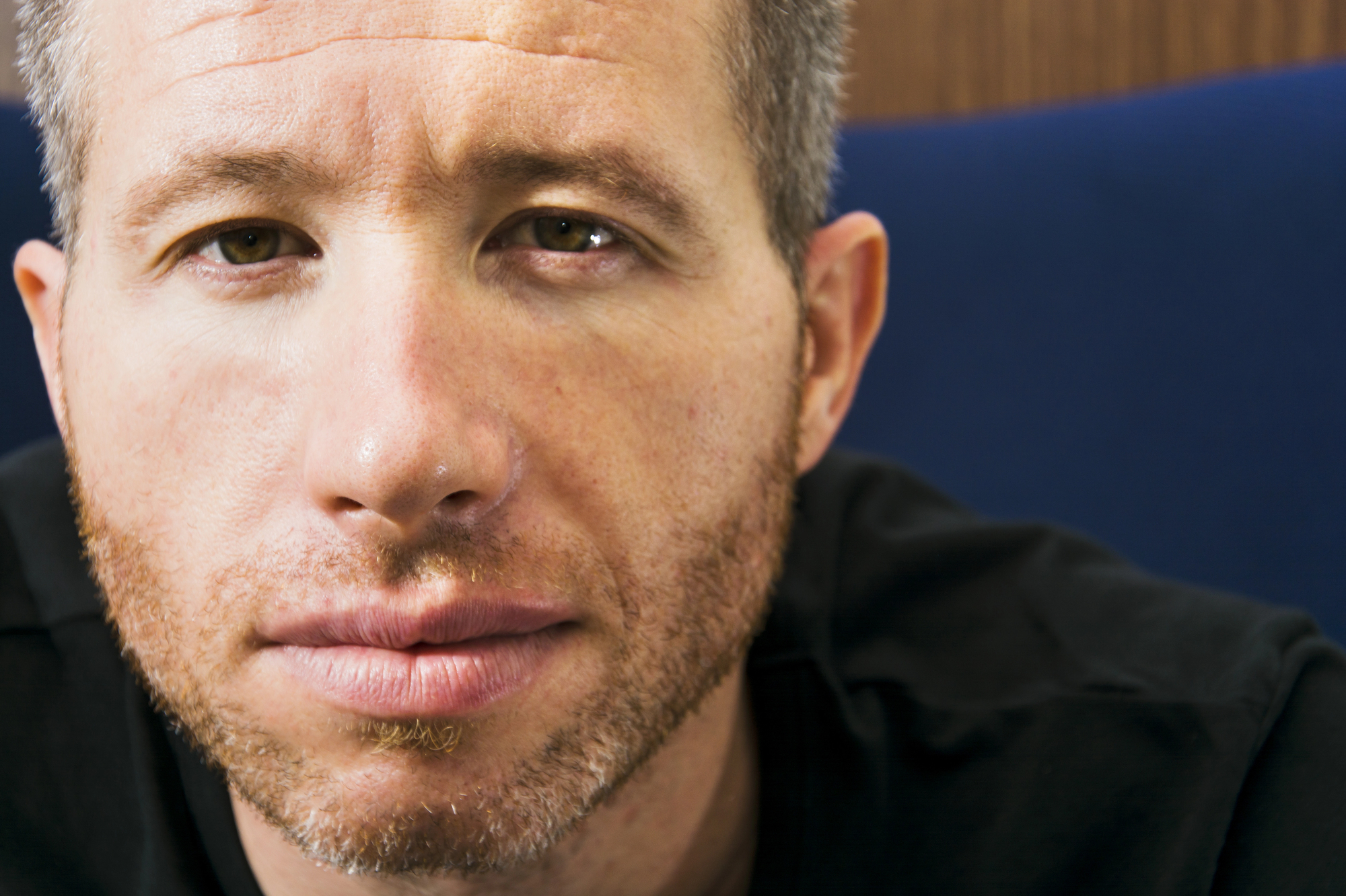 Close-up image of a man with short, graying hair and a light beard and mustache. He is wearing a black shirt and looking directly at the camera against a blurred background, which appears to be a mix of blue and wood tones.