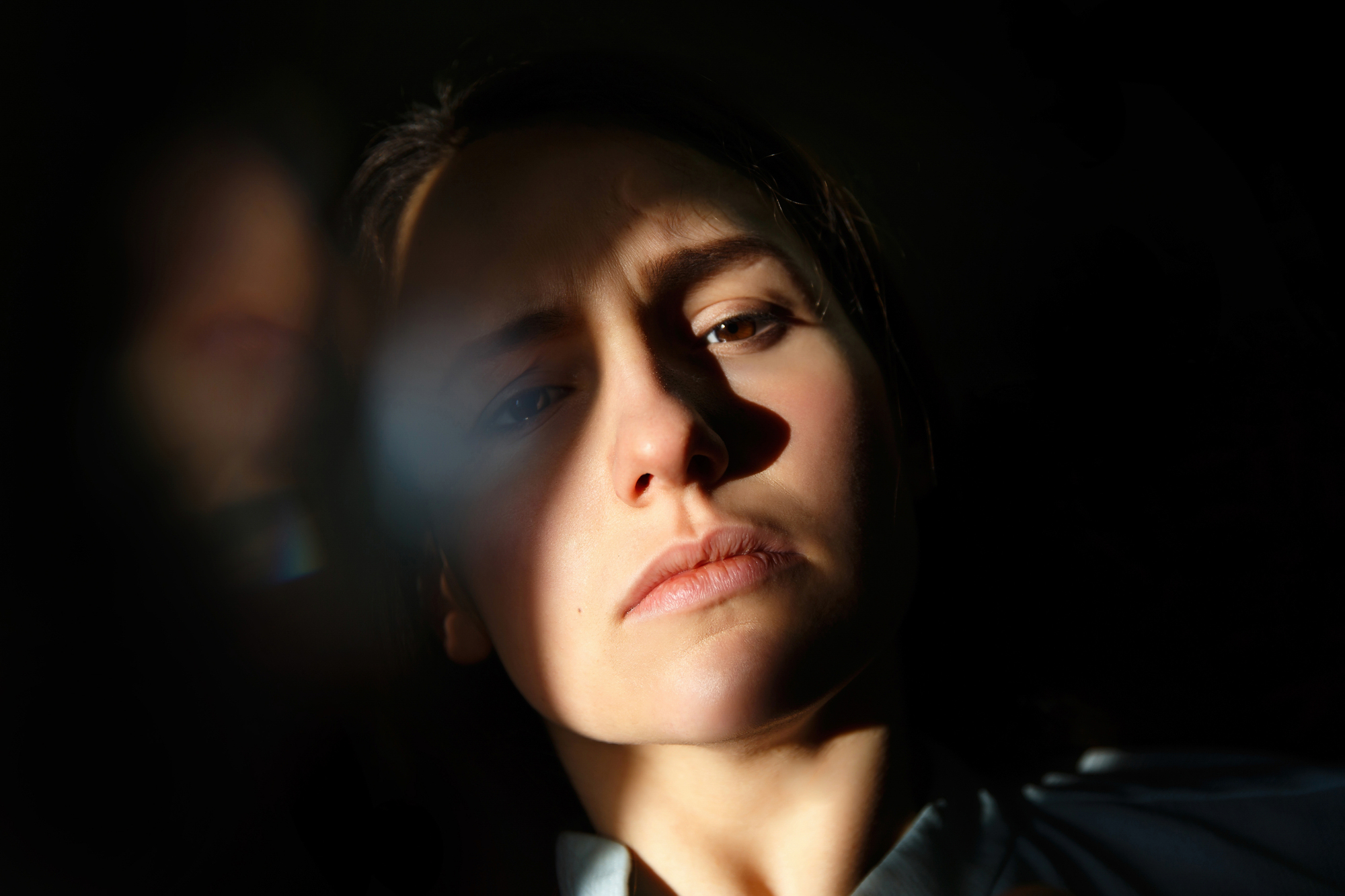 A close-up photo of a person with long hair, partially illuminated by soft light. The light casts shadows across their face, highlighting one cheek, part of their forehead, and lips. The background is dark, creating a dramatic and introspective mood.