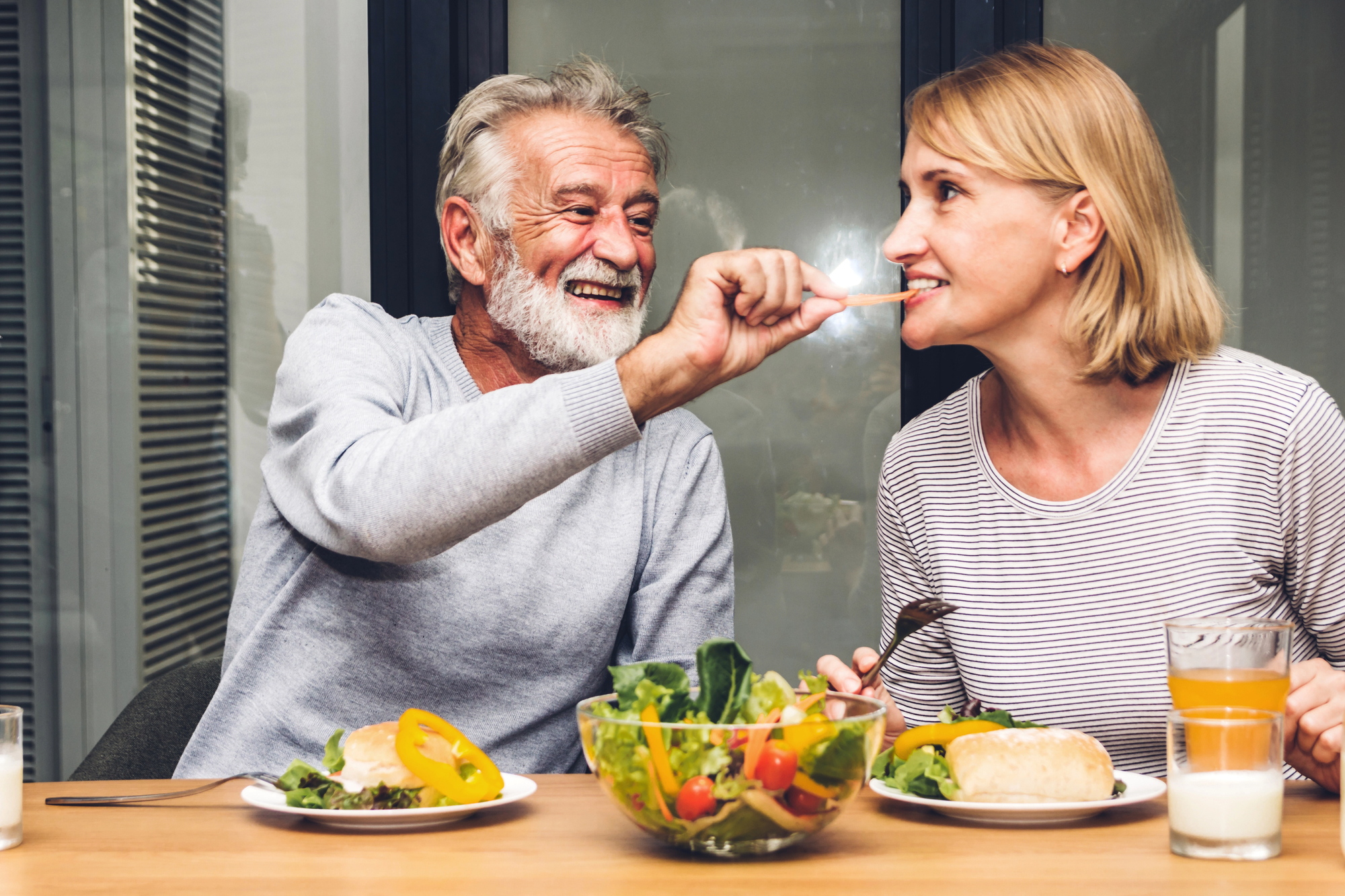 An elderly man playfully feeds a smiling woman a vegetable stick at a dining table. The table is set with plates of food, a bowl of salad, a glass of orange juice, and a glass of milk. They appear to be enjoying their meal and each other's company.