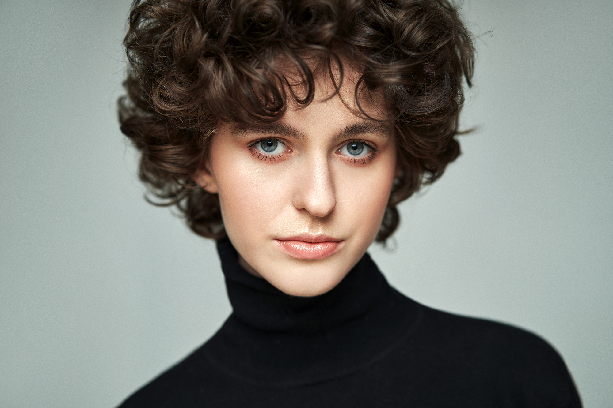 A person with curly brown hair and blue eyes is wearing a black turtleneck. They are looking directly at the camera with a neutral expression against a plain gray background.