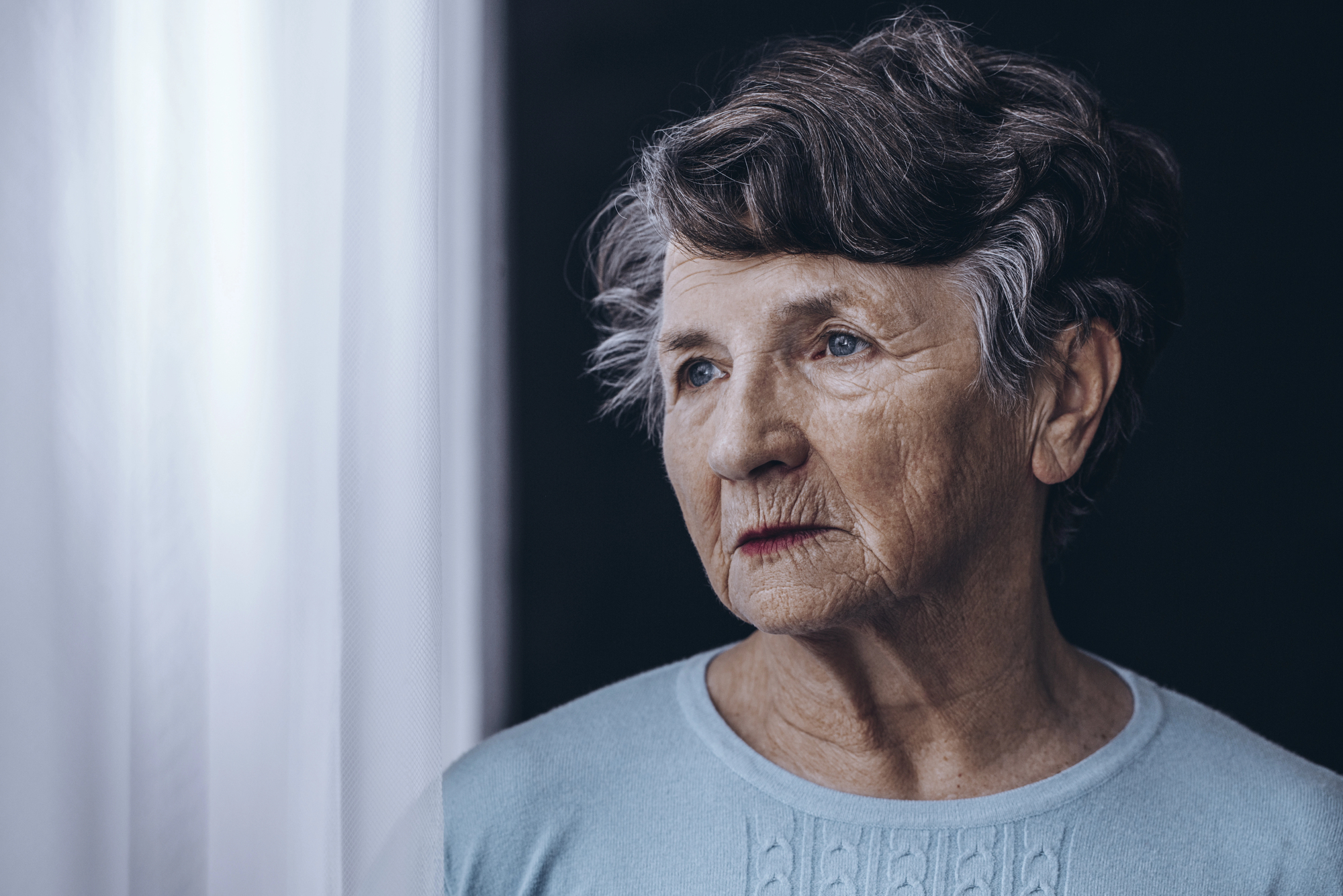 An elderly woman with short, wavy gray hair gazes thoughtfully out a window. She is wearing a light blue sweater, and the soft light from the window illuminates her face, highlighting her thoughtful expression and the wrinkles on her skin.