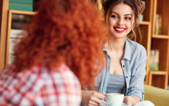 A woman with a bright smile, wearing a gray shirt, holds a cup of coffee while looking at another person with curly red hair. They are in a cozy indoor setting with bookshelves in the background.