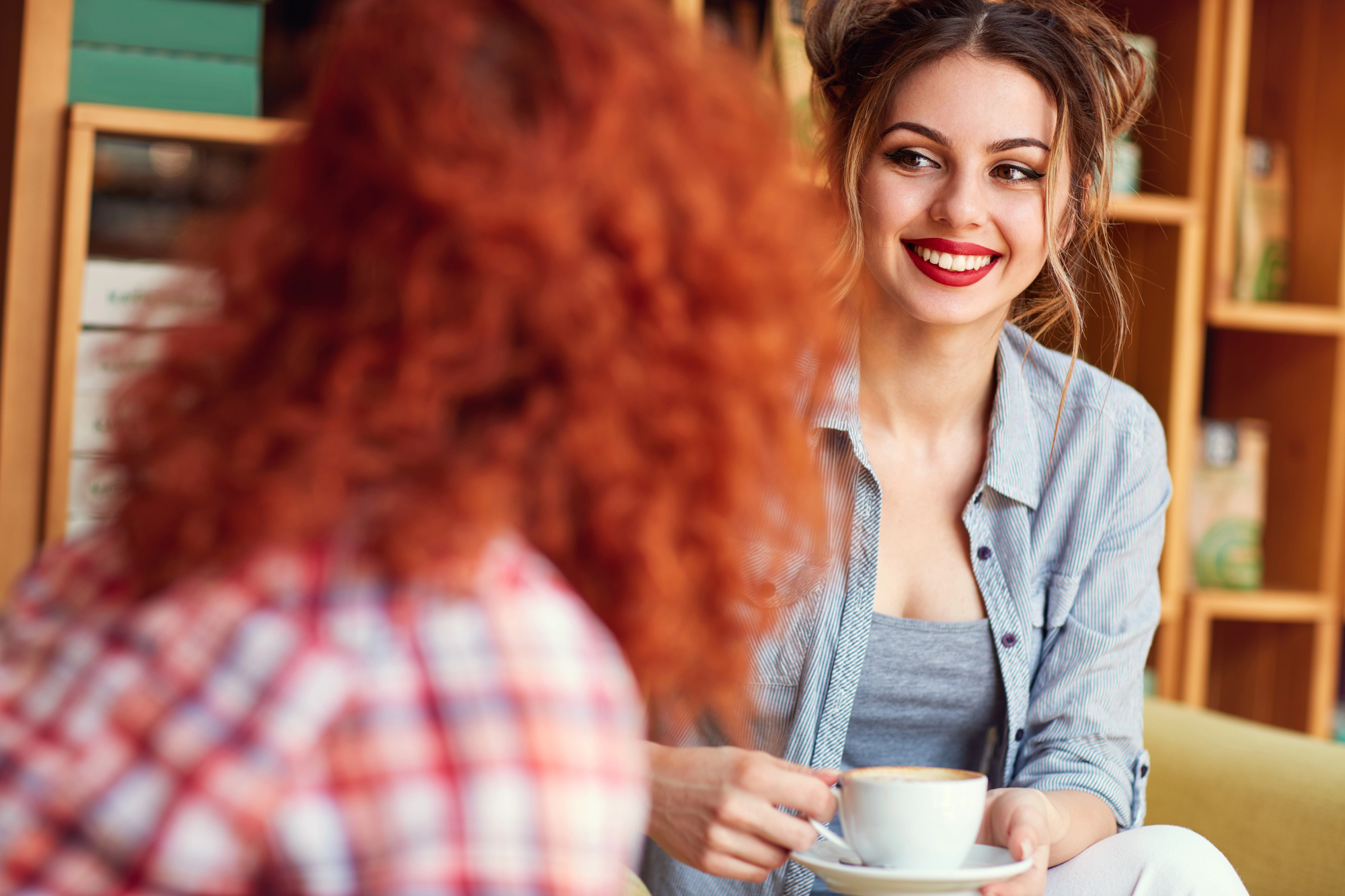 A woman with a bright smile, wearing a gray shirt, holds a cup of coffee while looking at another person with curly red hair. They are in a cozy indoor setting with bookshelves in the background.