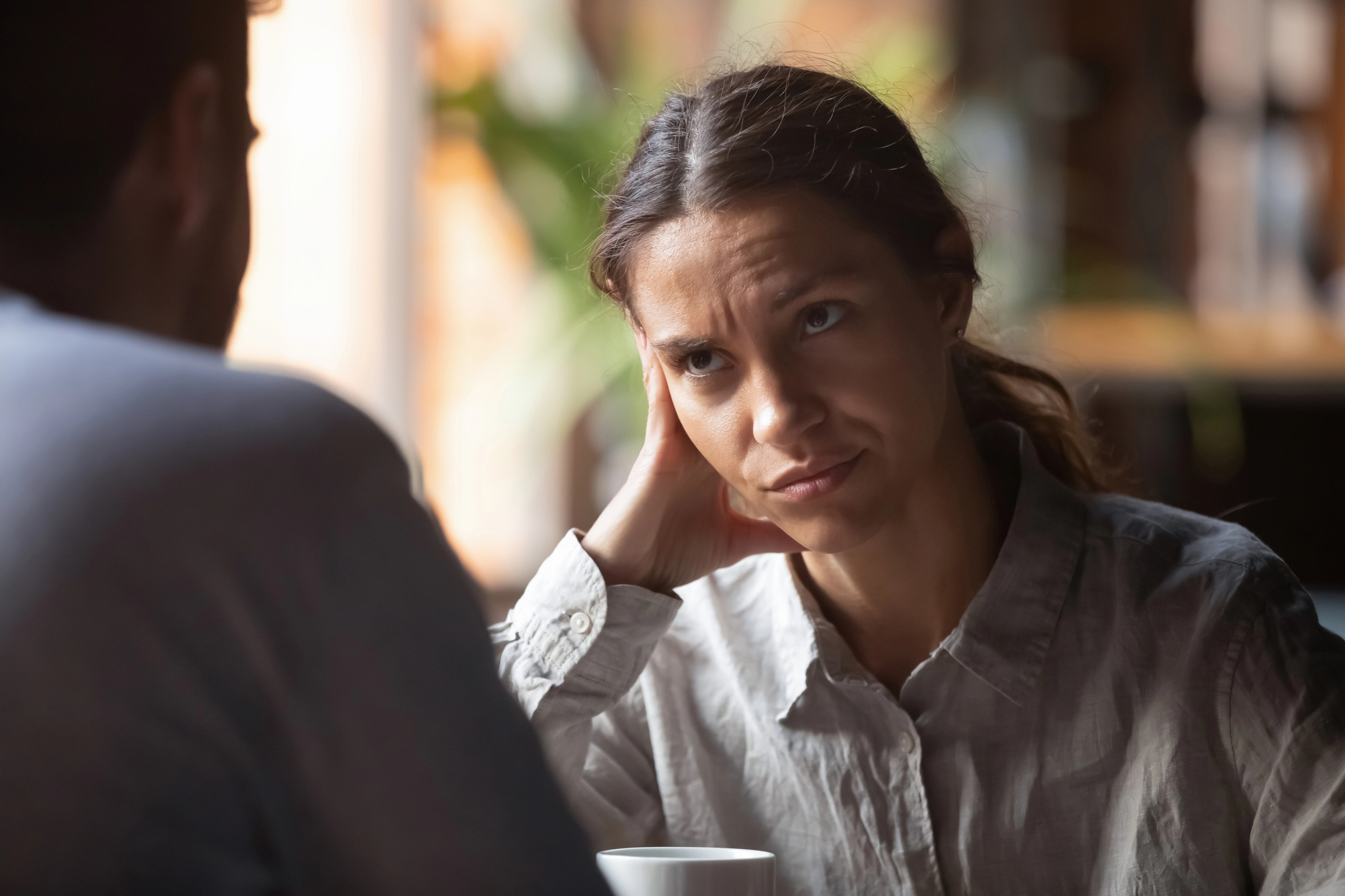 A woman with a skeptical expression rests her head on her hand while listening to someone. She is wearing a light-colored shirt and sitting indoors in front of a blurred background with a plant. A coffee cup sits on the table in front of her.