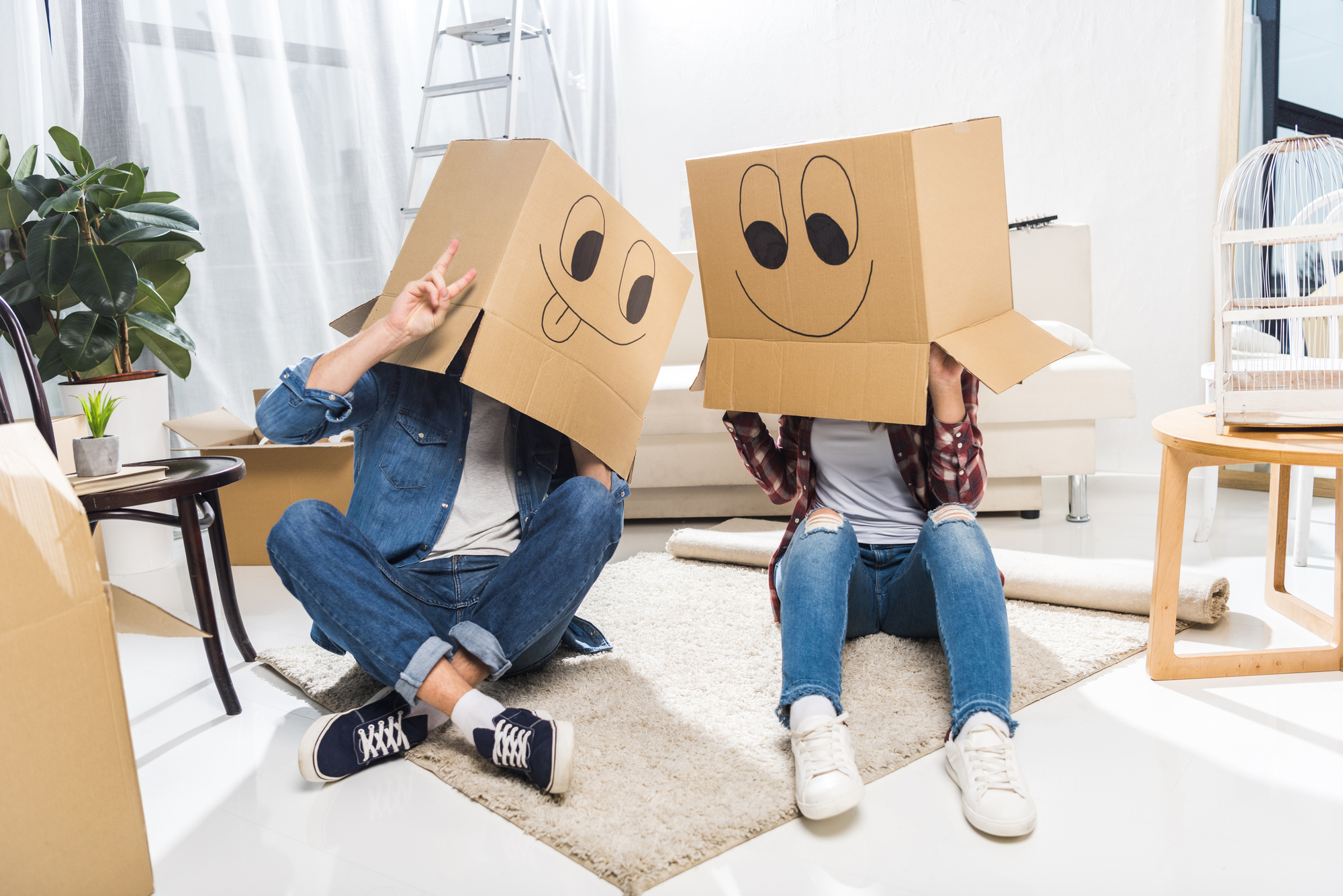 Two people sitting on a white rug, each wearing a cardboard box with cartoon faces drawn on them. They are inside a room with a white couch, a ladder, and plants in the background. One person has their legs crossed and is giving a peace sign.