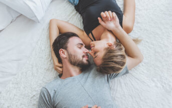 A man and woman are lying on a white, plush bedspread, facing each other affectionately. The man is wearing a gray shirt and the woman is wearing a black tank top. They are close, with their faces almost touching, and smiling softly.