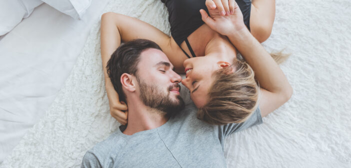 A man and woman are lying on a white, plush bedspread, facing each other affectionately. The man is wearing a gray shirt and the woman is wearing a black tank top. They are close, with their faces almost touching, and smiling softly.