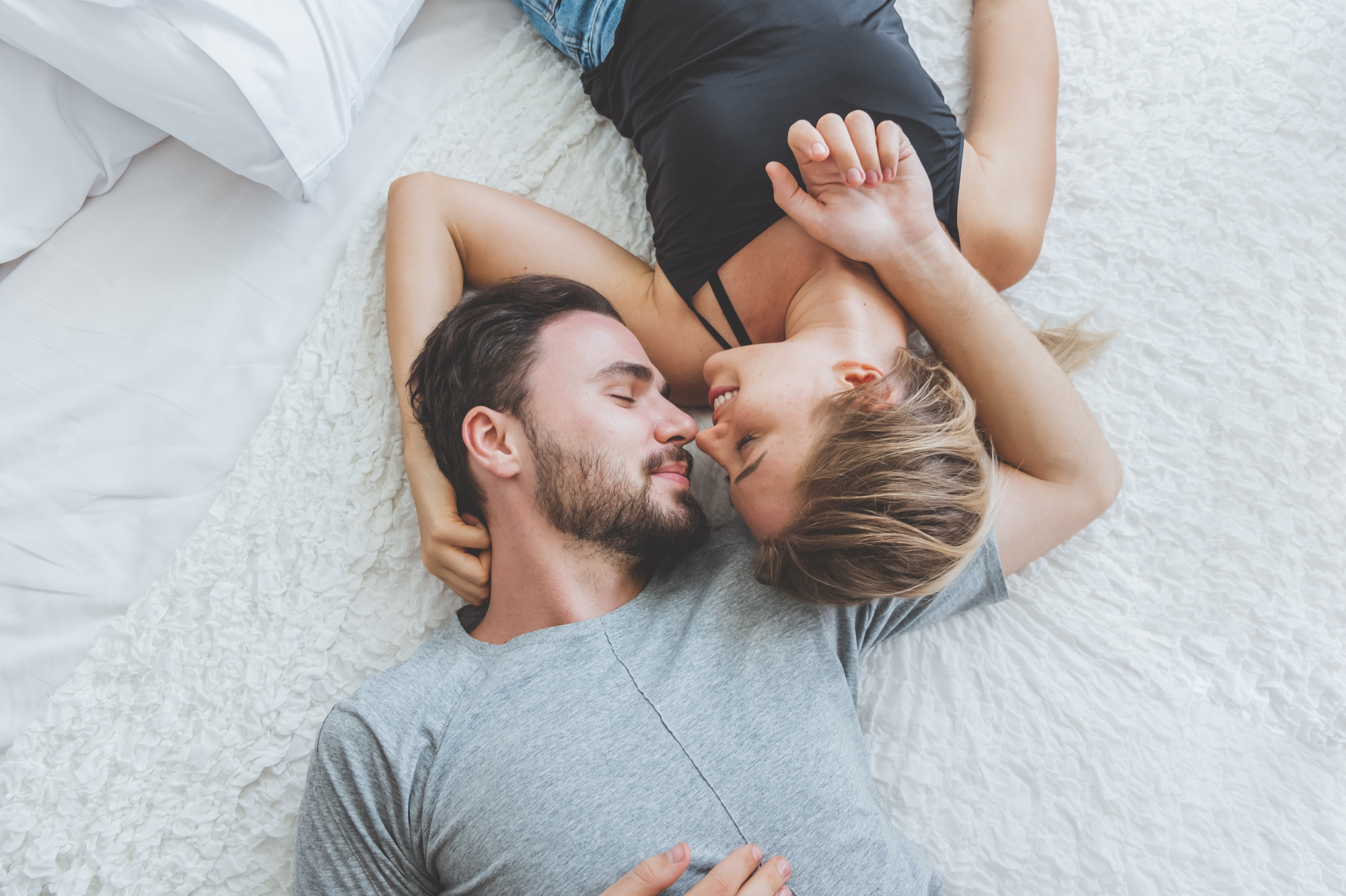 A man and woman are lying on a white, plush bedspread, facing each other affectionately. The man is wearing a gray shirt and the woman is wearing a black tank top. They are close, with their faces almost touching, and smiling softly.