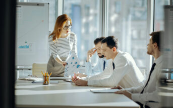 A woman with red hair in glasses and a striped shirt is standing and pointing at a document in a conference room with three seated men, all in business attire. A whiteboard with graphs and office supplies are visible in the background.