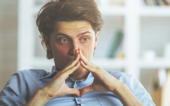 A person with brown hair sits indoors, looking thoughtful. They are resting their chin on their interlocked fingers. The background is blurry, with what appears to be bookshelves and various objects. The person is wearing a light blue button-up shirt.