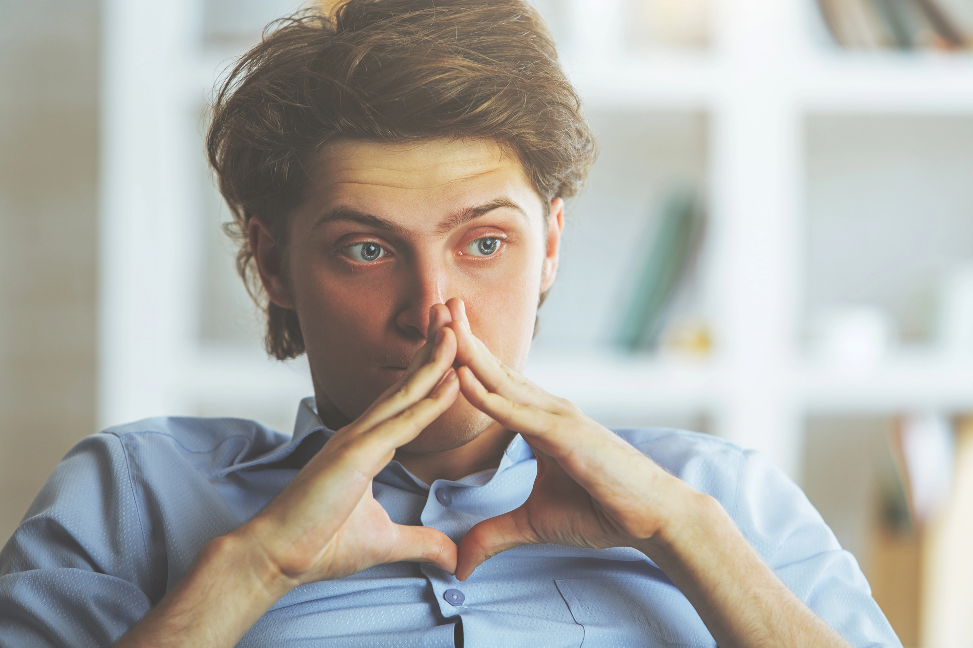 A person with brown hair sits indoors, looking thoughtful. They are resting their chin on their interlocked fingers. The background is blurry, with what appears to be bookshelves and various objects. The person is wearing a light blue button-up shirt.