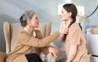 An elderly woman with gray hair and a younger woman with brown hair sit facing each other in a well-lit room. The elderly woman tenderly adjusts the younger woman's hair while they both smile. A beige armchair and modern decor are visible in the background.