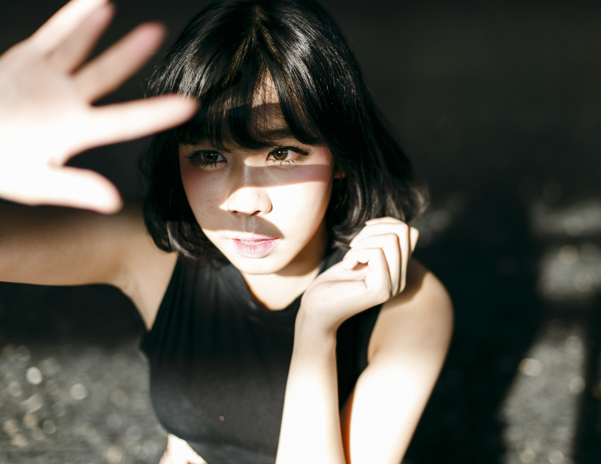 A person with short black hair and a black sleeveless top is shielding their face from the sunlight with one hand. Shadows and light play across their face, creating a dramatic effect. The background is out of focus and dark.