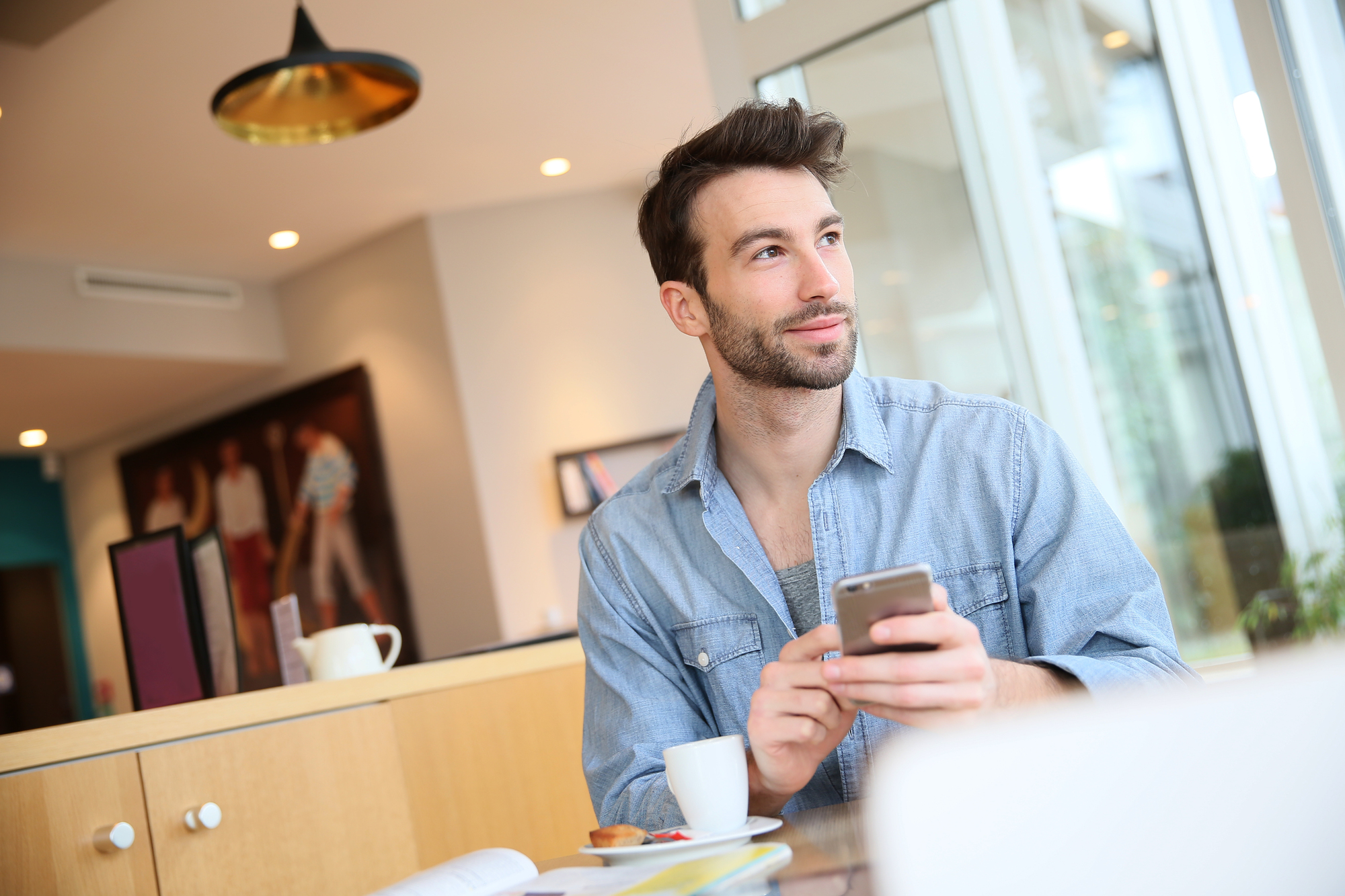 A man with short brown hair and a beard sits at a table in a modern cafe, holding a smartphone and looking thoughtfully out the window. He is wearing a light denim shirt, and there is a coffee cup, saucer, and an open book on the table.