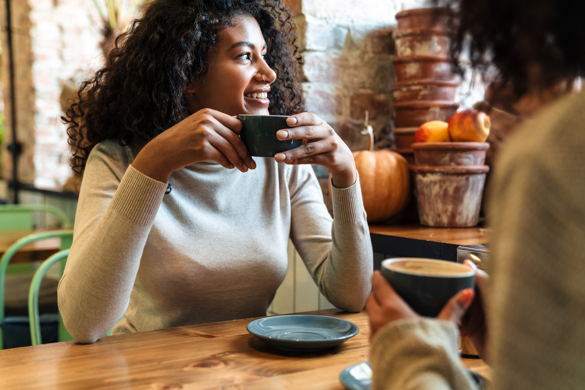 A woman with curly hair, wearing a beige sweater, is sitting at a wooden table and enjoying a cup of coffee. She is smiling and looking to the side. Another person, partially visible, is holding a cup nearby. The background includes pumpkins and pottery.