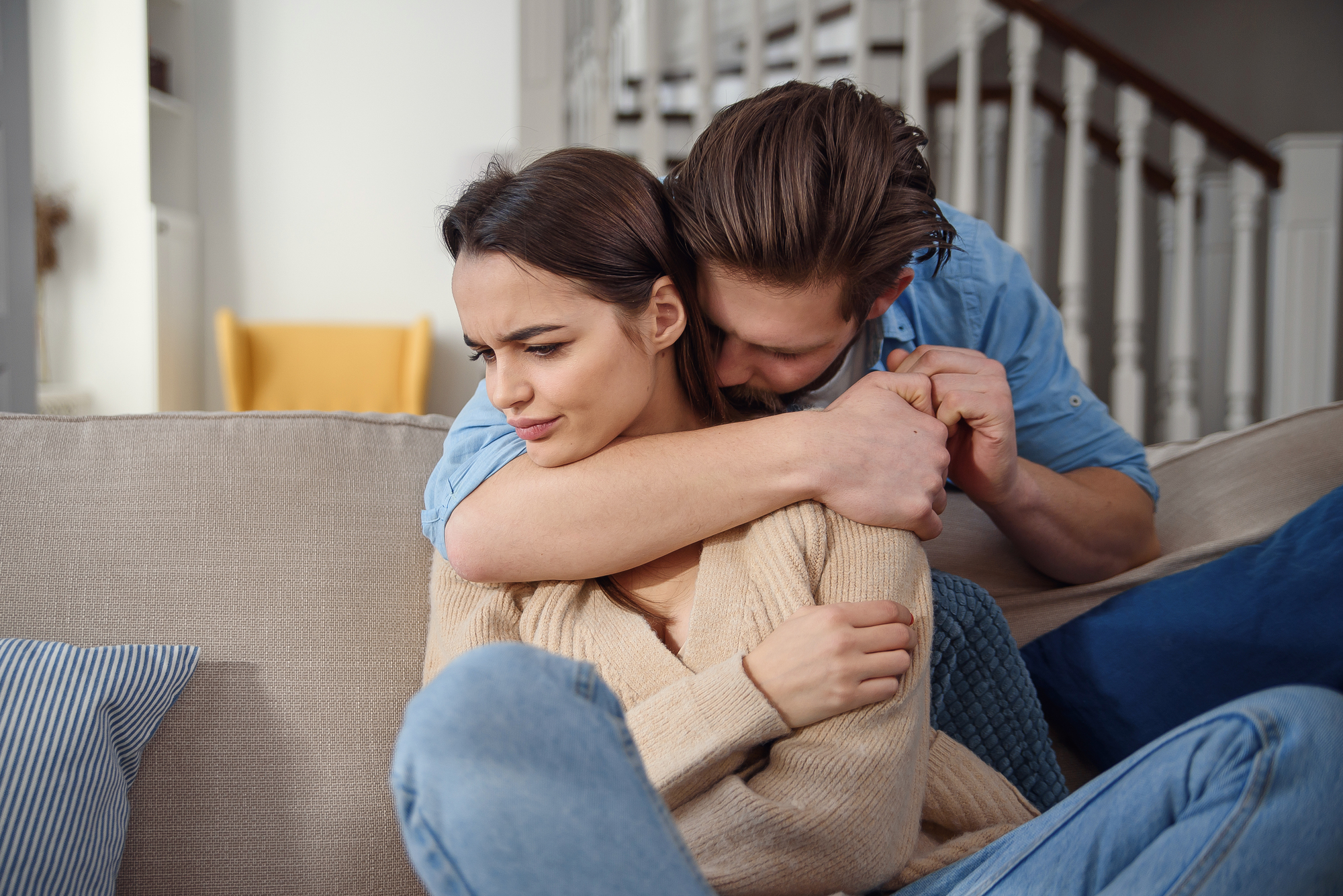 A woman sits on a couch, appearing upset and looking away, while a man sits behind her, wrapping his arms around her shoulders in an attempt to comfort her. She has her arms crossed, and they are in a cozy home setting with a staircase in the background.