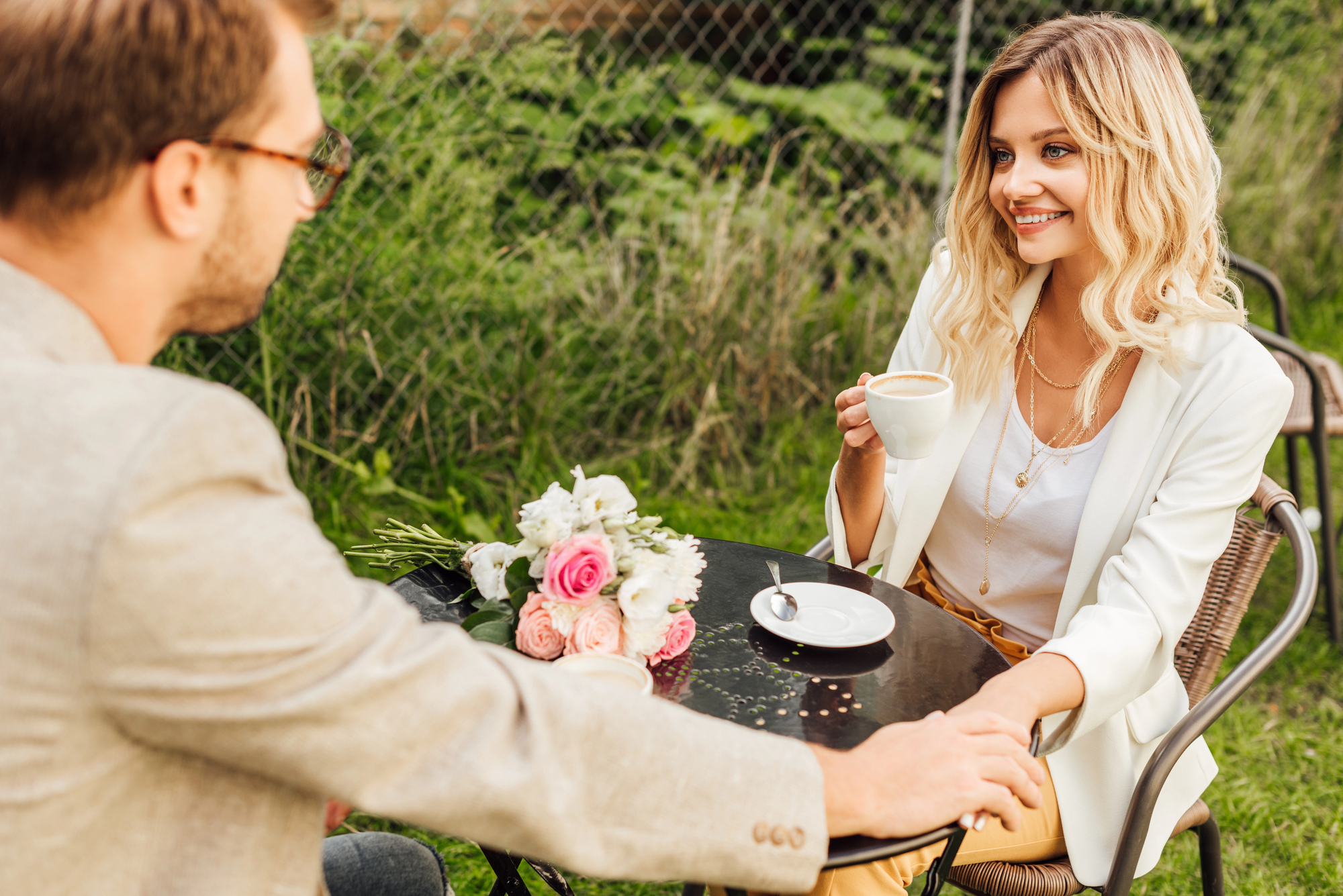 A couple seated at an outdoor café table with the woman holding a coffee cup and smiling at the man. The table has a bouquet of flowers and a saucer. They both appear to be enjoying a pleasant conversation amidst a lush, green background.