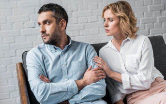 A man with short brown hair and a beard is sitting on a couch with his arms crossed, looking away to his left. A woman with blonde hair is sitting beside him, gently holding his arm and looking at him with concern. They are in a room with a white brick wall.