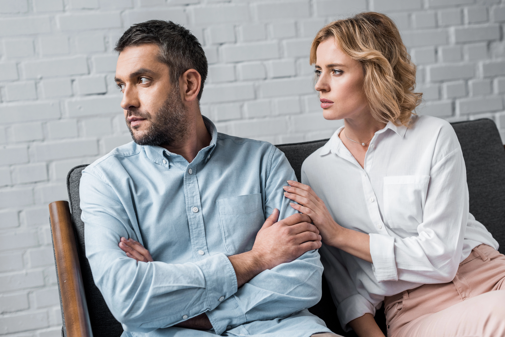 A man with short brown hair and a beard is sitting on a couch with his arms crossed, looking away to his left. A woman with blonde hair is sitting beside him, gently holding his arm and looking at him with concern. They are in a room with a white brick wall.