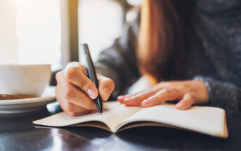 A person sits at a table, writing in a notebook with a pen. They are wearing a gray sweater, and a white cup is visible next to them on the table. Sunlight streams through a window in the background, creating a warm ambiance. The focus is on the person's hand and the notebook.