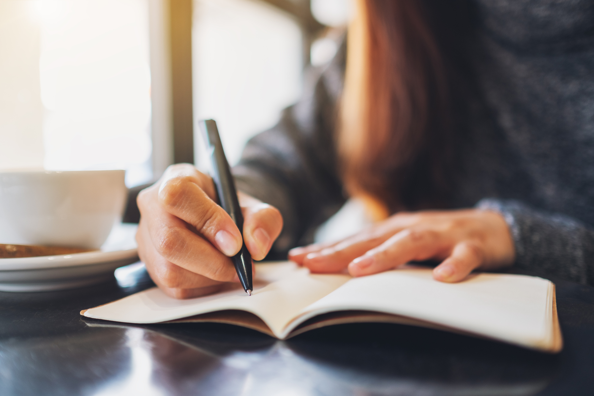 A person sits at a table, writing in a notebook with a pen. They are wearing a gray sweater, and a white cup is visible next to them on the table. Sunlight streams through a window in the background, creating a warm ambiance. The focus is on the person's hand and the notebook.