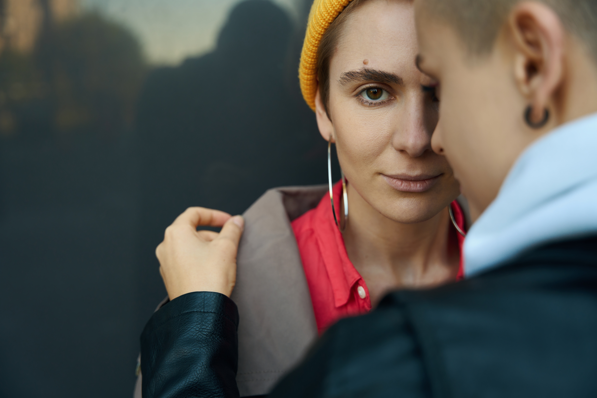 A person wearing a yellow beanie and red shirt gazes intently at the camera. Another person, with their back to the camera, gently holds the first person's collar. They both have short hair, and the setting is a dimly lit area, adding a pensive mood to the scene.