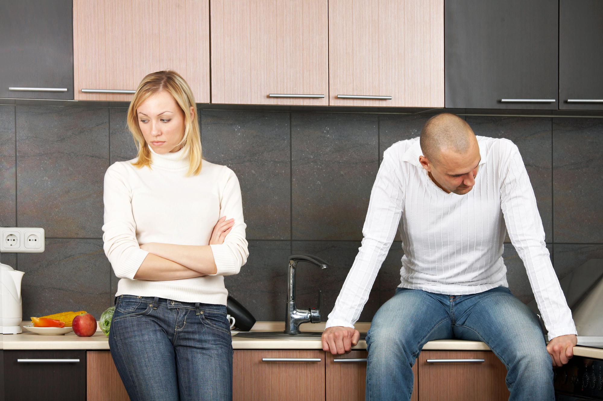 A woman and a man are in a kitchen. The woman stands with her arms crossed, looking away. The man sits on the counter, looking down. Both appear serious. The kitchen has wooden cabinets and a countertop with a fruit bowl.