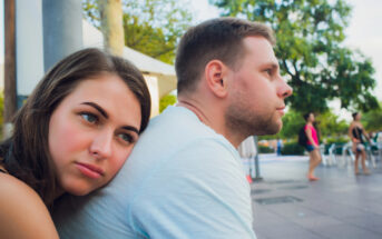 A young woman rests her head on a man's shoulder while both sit outside, with trees and people in casual attire in the background. The woman looks thoughtful, while the man appears to be looking ahead. The atmosphere is relaxed and summery.