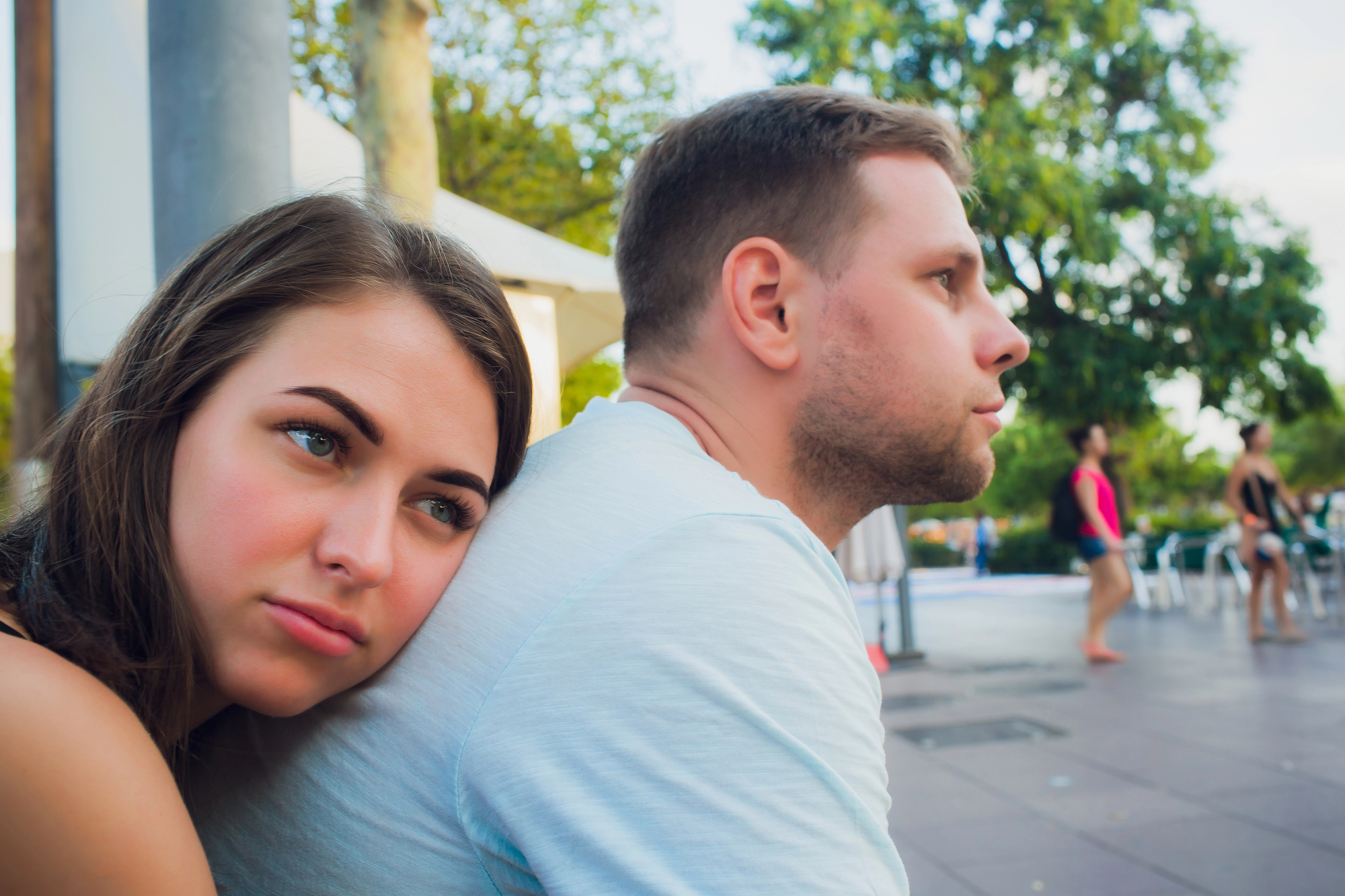 A young woman rests her head on a man's shoulder while both sit outside, with trees and people in casual attire in the background. The woman looks thoughtful, while the man appears to be looking ahead. The atmosphere is relaxed and summery.