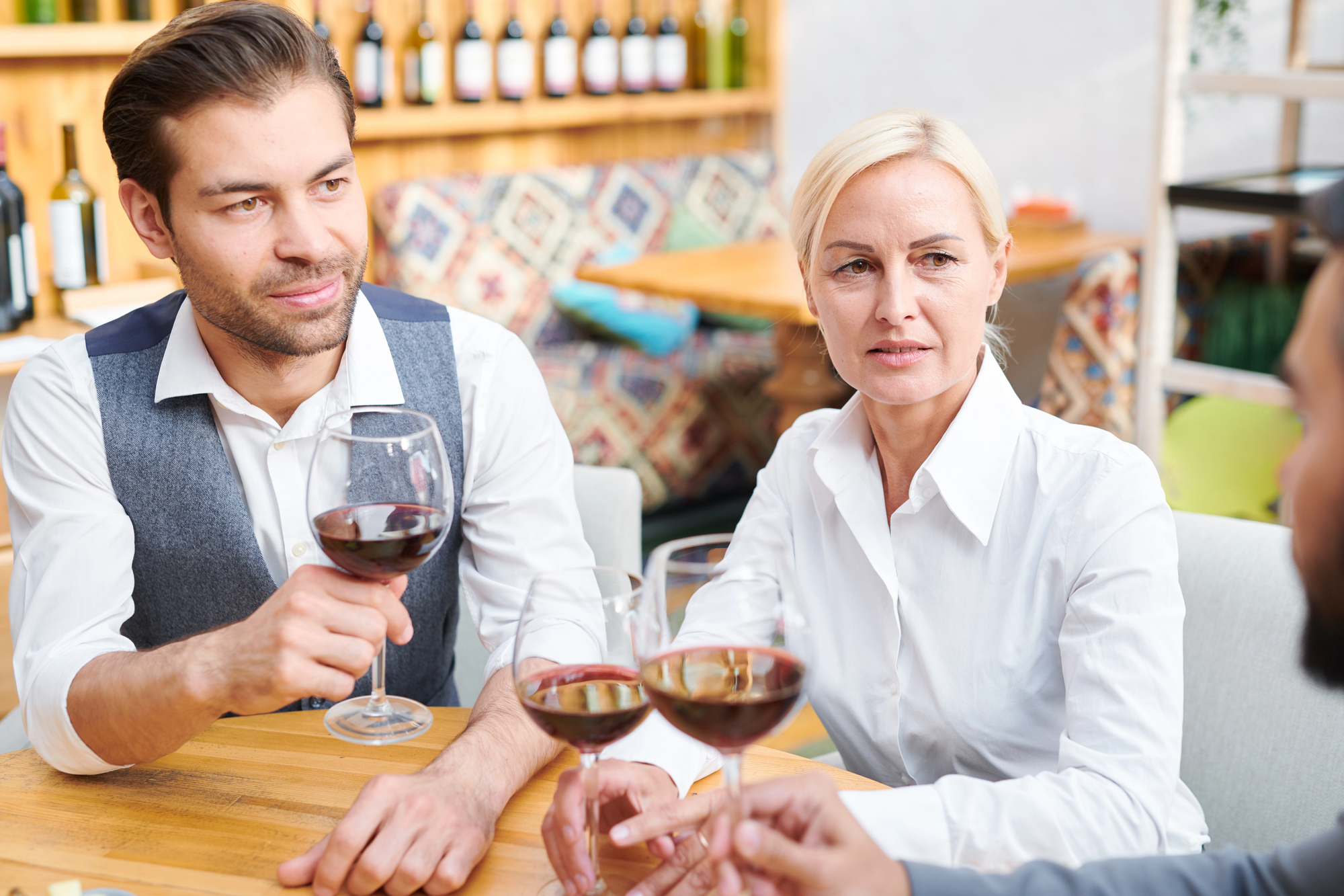 Two people are sitting at a table in a restaurant, engaging in conversation and holding glasses of red wine. A third person, partly visible, is joining them in a toast. Shelves with wine bottles are in the background, and the setting is cozy and casual.