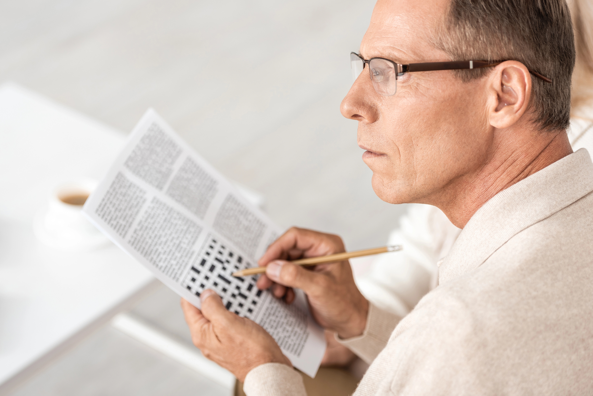 A man wearing glasses and a beige sweater is sitting at a table, holding a pencil and working on a crossword puzzle in a newspaper. A cup of coffee is partially visible on the table in the background.