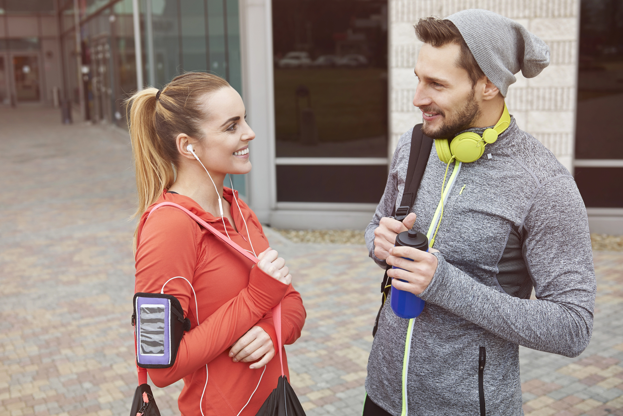 Two people in athletic wear stand outdoors near a building, smiling and talking. One wears a red top with a phone armband and earphones, holding a black gym bag. The other wears a gray top, beanie, and yellow headphones, holding a blue water bottle.