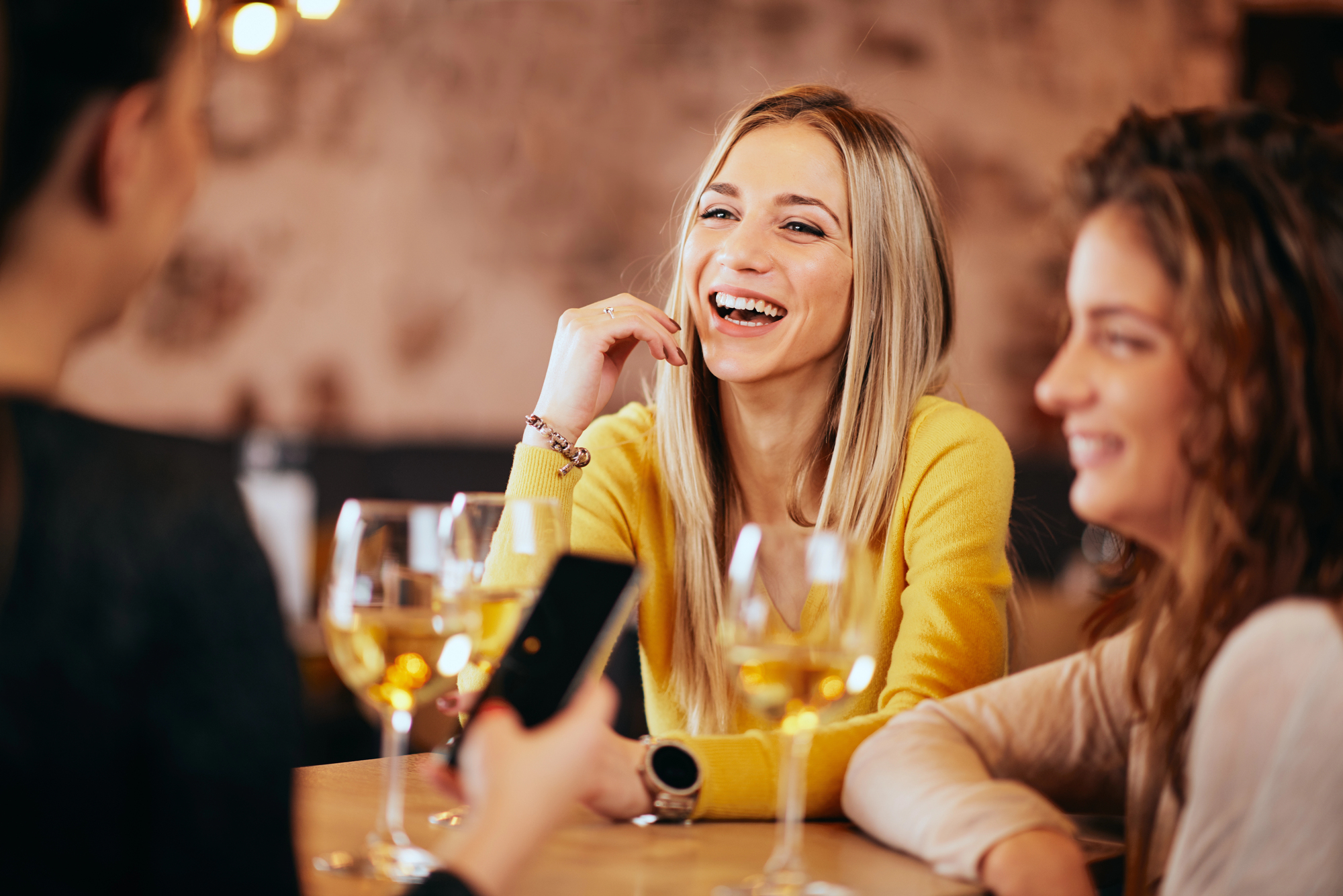 Three women enjoying a conversation in a cozy setting, with wine glasses on the table in front of them. One woman, in a yellow sweater, is laughing, while the other two are smiling. Warm lighting creates a relaxed atmosphere.