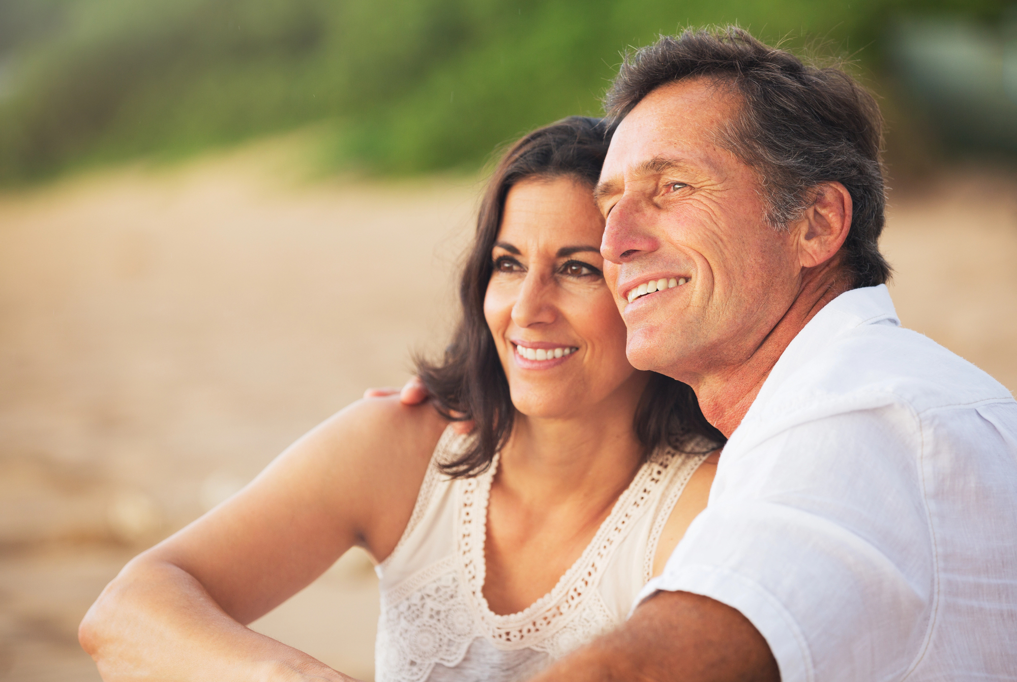 A middle-aged couple sits closely together on a beach, smiling and looking into the distance. The woman has dark hair and is wearing a white sleeveless top, while the man, also in white, has graying hair. The background is blurred with greenery and sand.