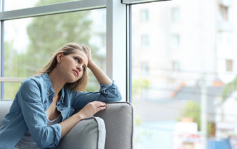 A young woman with long hair sits on a couch beside a large window, resting her head on one hand. She is wearing a denim shirt over a white top and appears to be deep in thought, gazing out the window at the blurry buildings and greenery outside.