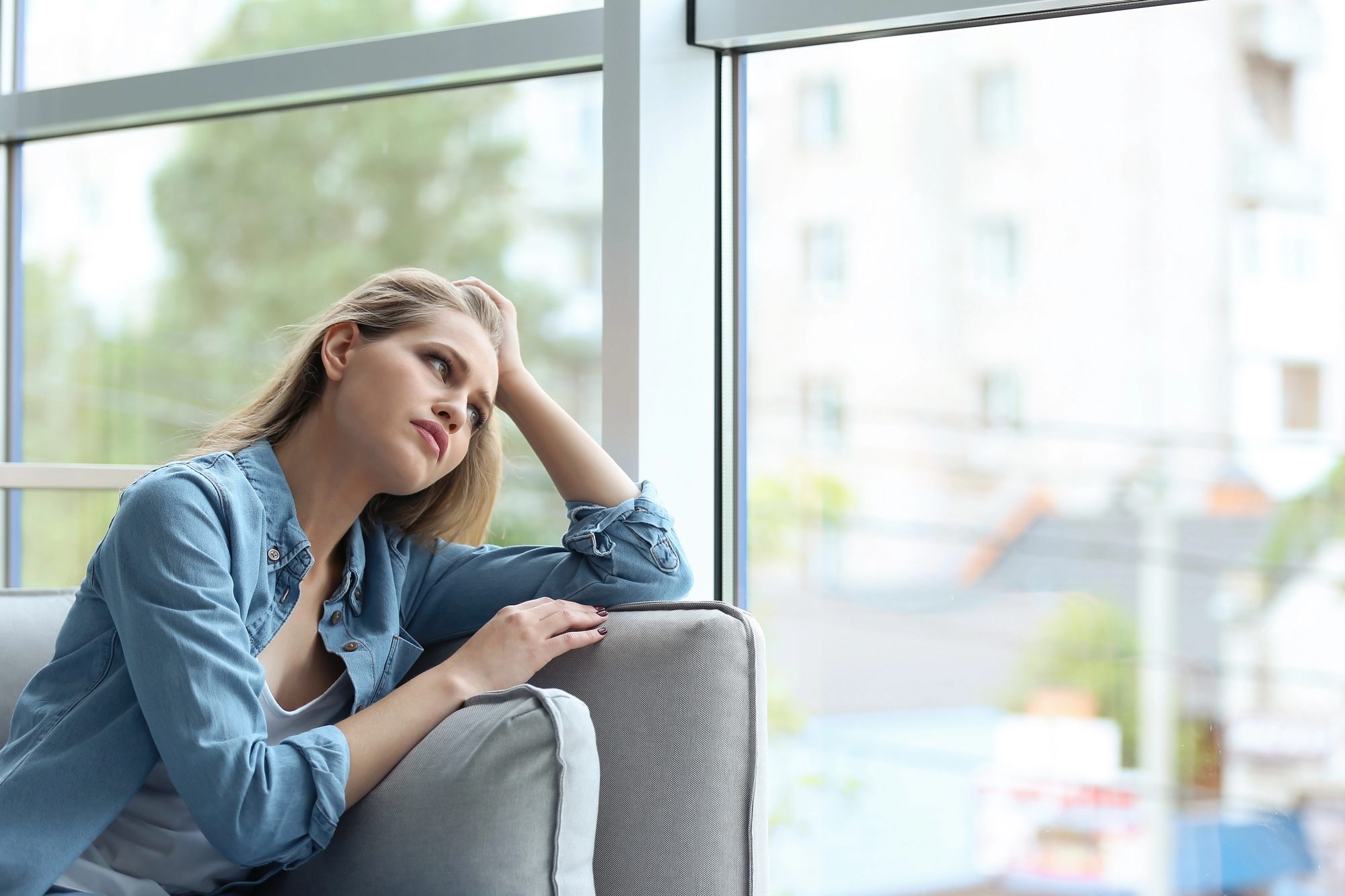 A young woman with long hair sits on a couch beside a large window, resting her head on one hand. She is wearing a denim shirt over a white top and appears to be deep in thought, gazing out the window at the blurry buildings and greenery outside.