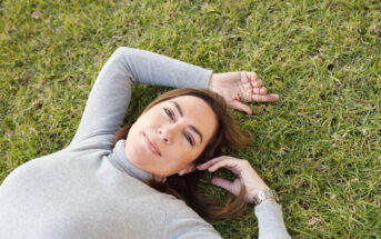 A woman lies on green grass with her hands relaxed above her head. She has a light gray long-sleeve shirt and a watch on her left wrist. Her eyes are open, and she has a peaceful expression on her face.