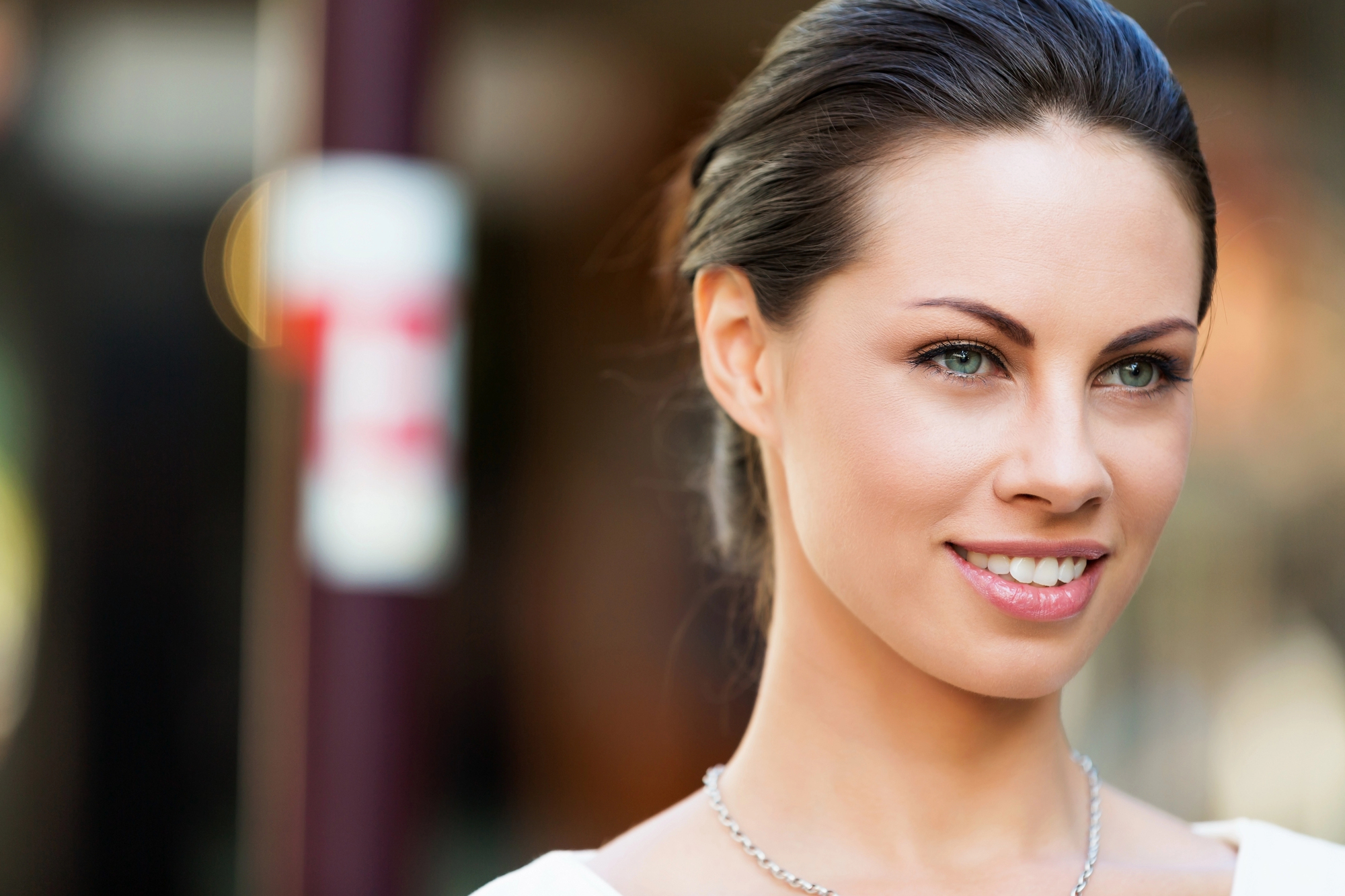 A woman with light skin and blue eyes, smiling, with her hair pulled back. She is wearing a silver necklace and a light top. The background is blurred but appears to be outdoors with light and dark areas.